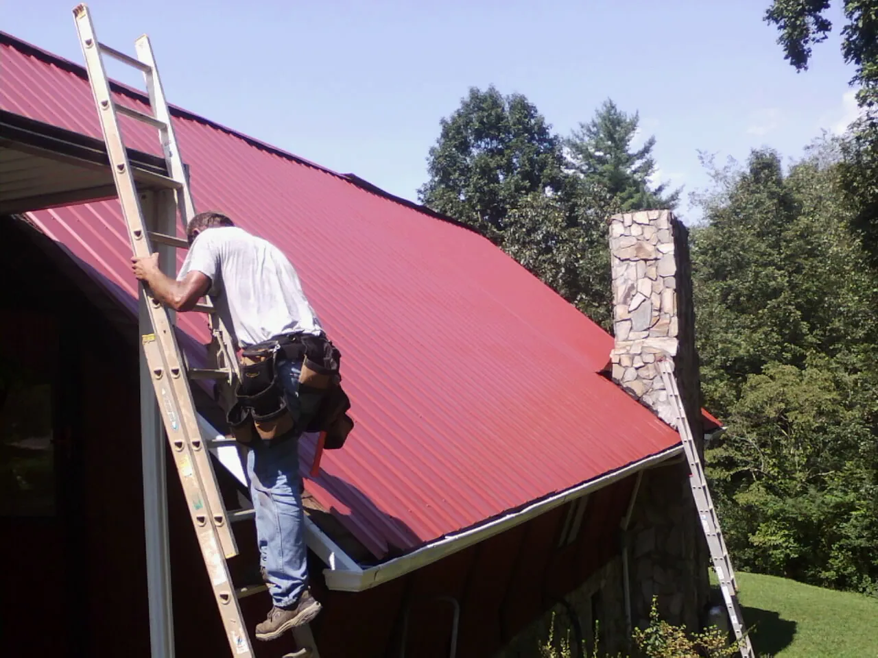 Roofer on a ladder beside a red metal roof and stone chimney — Soffit & Fascia Repair by Roofing Brothers in Clearlake